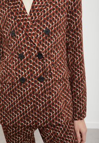 Punainen ja ruskea kuvioitu puku, jossa on kuusikulmaisia muotoja, kaksirivinen malli, mustat napit ja taskuyksityiskohdat. Raskaskankainen materiaali.