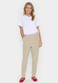 Beige skräddarsydda byxor med veck, bärs tillsammans med en vit t-shirt i V-form. Röda platta skor och en neutral bakgrund fulländar outfiten.