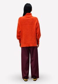 Persoon met lang zwart haar, gekleed in een oranje coltrui, wijde paarse broek en geel-brown schoenen, staande met de rug naar de kijker gekeerd.