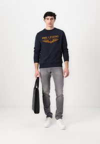 Marineblauwe sweatshirt met oranje tekst en vleugelafbeelding, grijze jeans, witte sneakers en een zwarte tas; casual outfit met moderne pasvorm.