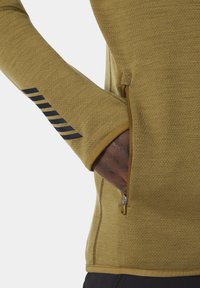 Długi rękaw, musztardowy top o fakturowanej tkaninie, z bocznym zamkiem błyskawicznym i czarnymi paskami na rękawie.