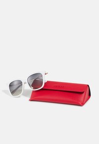 White framed sunglasses with dark lenses resting next to a closed red textured rectangular case embossed with "GUESS" on a white background.