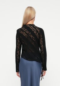 Schwarzes Spitzentop mit langen Ärmeln und strukturiertem Muster, das seitliche Bindedetails und gewellte Kanten aufweist, von hinten an einem Modell präsentiert.