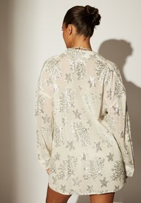Cubierta transparente de manga larga en color beige claro con patrones de estrellas de mar y corales plateados, que presenta un corte holgado y un cierre con lazo en el cuello.