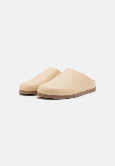 Sabots beiges avec une finition lisse et mate ; design à enfiler, bout arrondi et semelle marron ; forme simple et moderne sans motifs visibles.