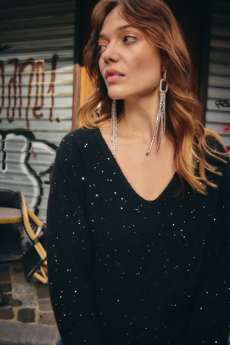 Juodas megztas megztinis su V formos iškirpte, papuoštas išmėtytais sidabriniais blizgučiais. Ilgi, žėrintys auskarai papildo įvaizdį, fone matomas grafičio piešinys.