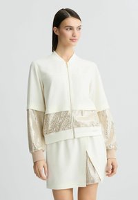 Cream bomber jacket with shiny gold sequin sleeves and matching hem, featuring a front zipper and ribbed cuffs. Short skirt with similar detailing.