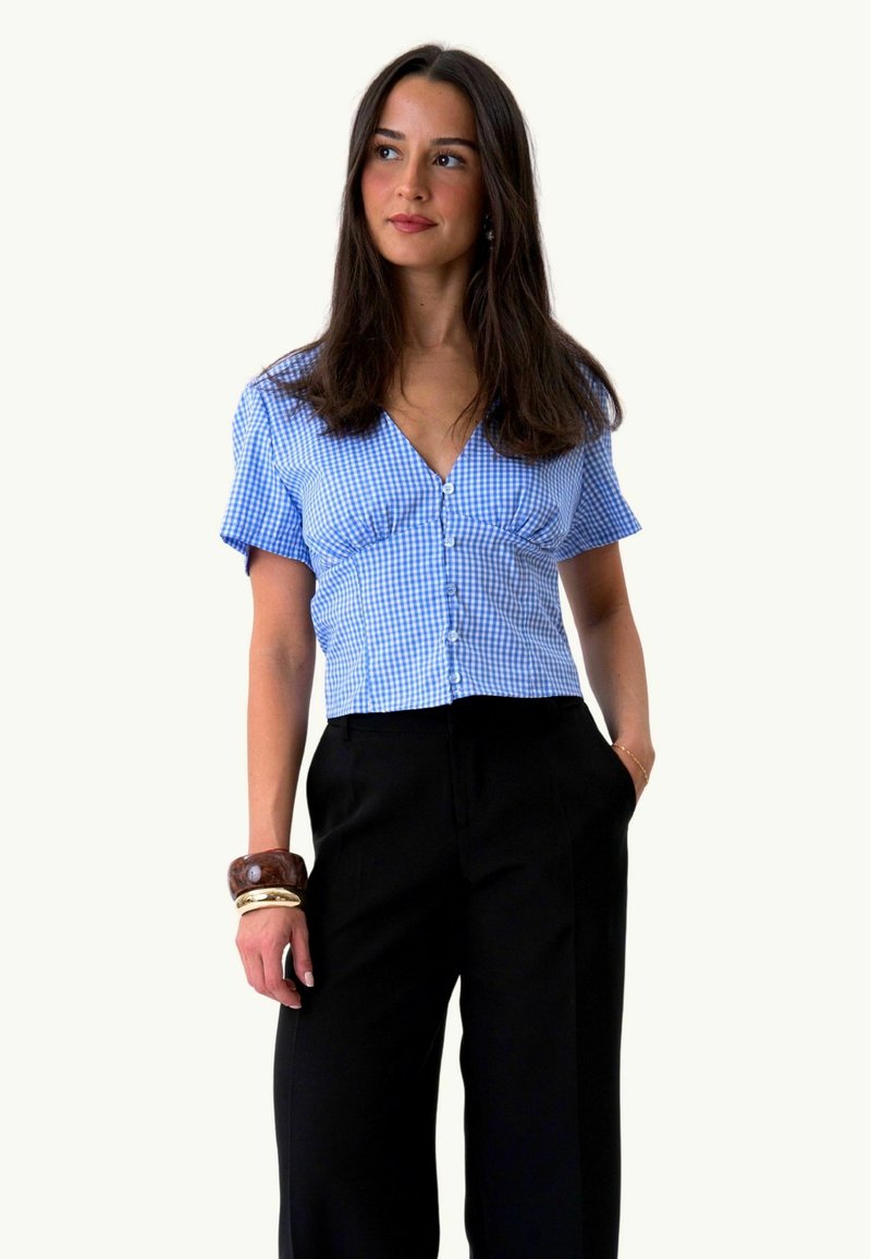 Jeune femme aux longs cheveux foncés portant un chemisier bleu à carreaux à manches courtes et un pantalon noir, avec des bracelets à son poignet gauche.