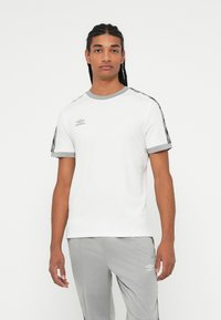 Ungdom med flettehår iført hvite og grå sportsklær med Umbro-logoer, stående mot en enkel hvit bakgrunn.