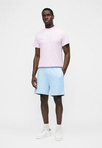 Άνδρας στέκεται φορώντας ανοιχτό μωβ μπλουζάκι, ανοιχτό μπλε σορτς, λευκές κάλτσες και λευκά αθλητικά παπούτσια σε απλό λευκό φόντο.