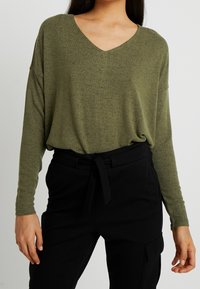 Top a maniche lunghe verde oliva con scollatura a V, realizzato in tessuto morbido e strutturato, abbinato a pantaloni neri a vita alta con cintura annodata.