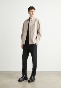 Beige button-up shirt with a single chest pocket, paired with black trousers and black shoes featuring a thicker sole and smooth texture.