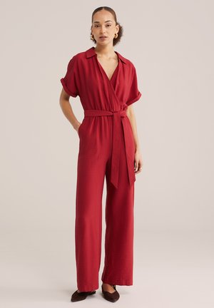 Vrouw draagt een rode jumpsuit met korte mouwen en riem, met brede pijpen, staat met één hand in de zak en een neutrale uitdrukking.