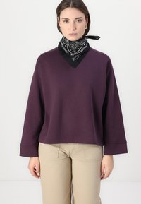 Felpa viola a scollo a V con maniche lunghe, abbinata a un bandana bianco e nero attorno al collo. Pantaloni beige visibili sotto.