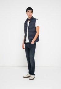 Tamsiai mėlynas pūstas liemenėlis su horizontaliomis siūlėmis, aukštu apykakliu, užsegama užtrauktuku; dėvima ant baltos marškinėlių palaidinės ir derinama su tamsiomis džinsais bei šviesiais sportbačiais.