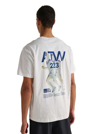 Mann trägt weißes T-Shirt mit blauem Text "ATW 223" und Grafik von zwei Läufern auf dem Rücken, stehend vor weißem Hintergrund.