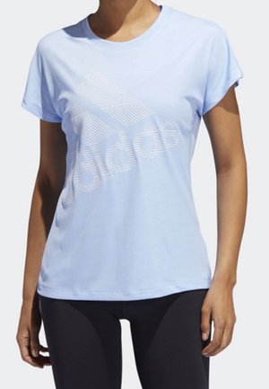 Femme portant un t-shirt Adidas bleu clair avec un grand logo rayé et des leggings noirs, debout devant un fond uni.