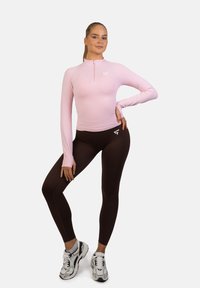 Jasnoróżowa bluzka z długim rękawem i zamkiem, w połączeniu z brązowymi legginsami. Wyraźne białe logo na bluzce i legginsach. Sportowe obuwie w odcieniach szarości i czerni.