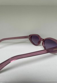 Roze doorschijnende zonnebril met een kattenoogvorm en een gradiënt paarse lenzen. Beschikt over smalle, gebogen armen met subtiele branding.