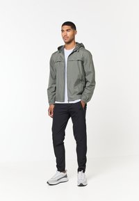 Letthurt, olivengrøn zip-jakke med hætte, elastiske håndled og to frontlommer, parret med sorte slim-fit bukser og grå sneakers.