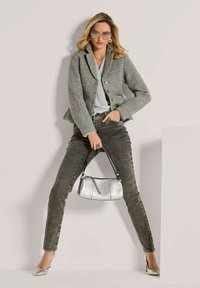 Vrouw in een grijze blazer, lichte trui, donkere skinny jeans, zilveren hakken en een zonnebril, met een zilveren handtas voor haar.