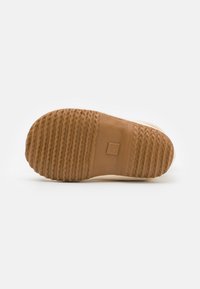 Semelle en caoutchouc avec une finition marron texturée, présentant des rainures pour une meilleure adhérence et un design plat. La partie claire près du talon est lisse.