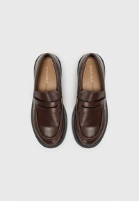 Mocassins en cuir marron à bout arrondi, dotés d'une lanière décorative et d'une semelle en caoutchouc noir. L'intérieur est doublé de beige avec le logo de la marque.
