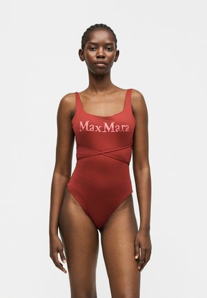 Kobieta ubrana w jednoczęściowy strój kąpielowy w kolorze rdzawo-czerwonym z cienkimi ramiączkami i logo "Max Mara" na wysokości klatki piersiowej, stojąca na tle jednolitego jasnego tła.