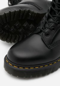 Botte noire en cuir à lacets avec un bout arrondi, une semelle en caoutchouc noire robuste et des détails de surpiqûres jaunes autour de la base.