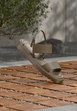 Brūna Calvin Klein sandale ar regulējamu potītes siksniņu, peld virs koka flīžu grīdas, ārā, ar nedaudz izplūdušu zaļumu un sienas fonu.