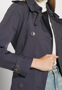 Marinblå trenchcoat i slätt tyg med hög krage, dubbelknäppt knäppning och justerbara ärmremmar. Har sidofickor.