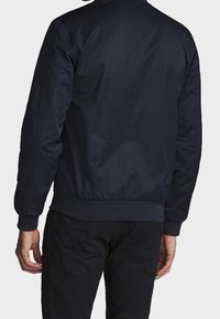 Chaqueta bomber azul marino con acabado liso y mate, puños y dobladillo acanalados, y detalle de costuras en los hombros. Diseño simple, sin logotipos visibles.