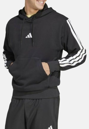 Person wearing a black Adidas hoodie with white stripes on sleeves and black pants with Adidas logo, hands in hoodie pocket, smiling looking right.