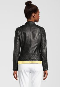 Zwarte leren jack met een gequiltte textuur op de schouders en de zoom. Hoge kraag en aansluitend ontwerp. Gelaagd over een geel shirt en witte broek.