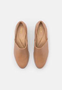 Chaussures en cuir sans lacets de couleur marron clair. Texture lisse, bout arrondi, et intérieur perforé pour la ventilation. Semelle intérieure de marque visible.