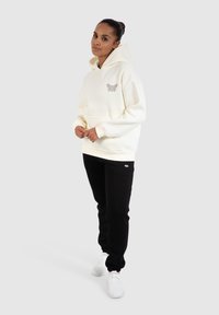 Cremefarbener Hoodie mit einer Fronttasche und gesticktem Design, kombiniert mit schwarzen Jogginghosen und weißen Turnschuhen vor einem grauen Hintergrund.