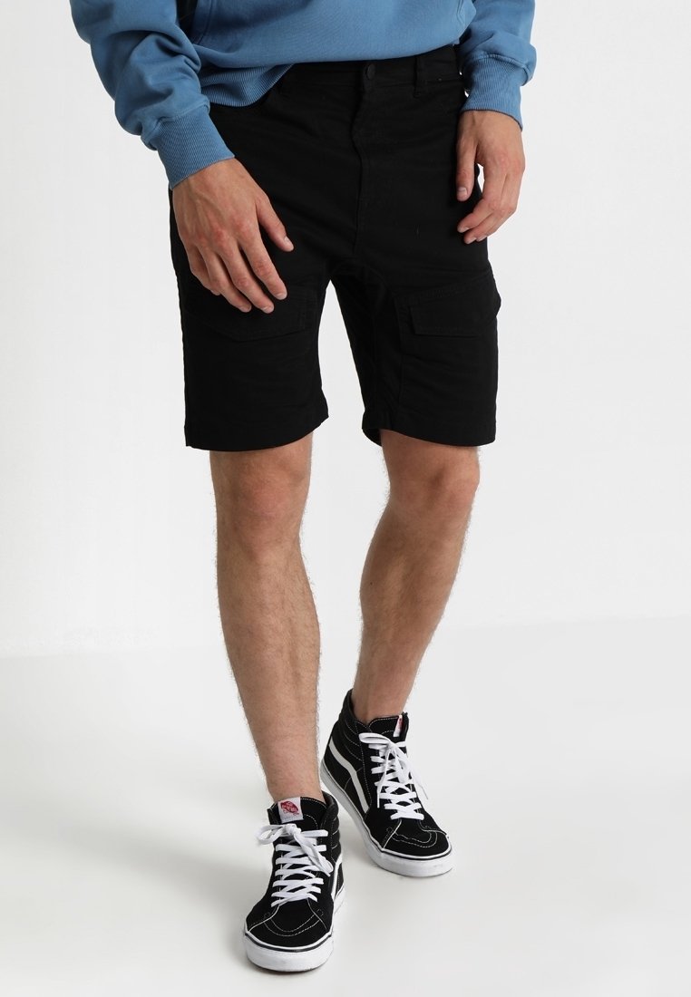 Homme portant un short cargo noir, des baskets montantes noires avec des lacets blancs et un sweat-shirt bleu, debout devant un fond uni.