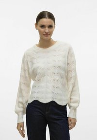 Aukštos kokybės megztinis off-white spalvos, su banguota raštų, raukiniais rankogaliais ir apvaliu hemu. Suderintas su tamsiai mėlynomis džinsomis.