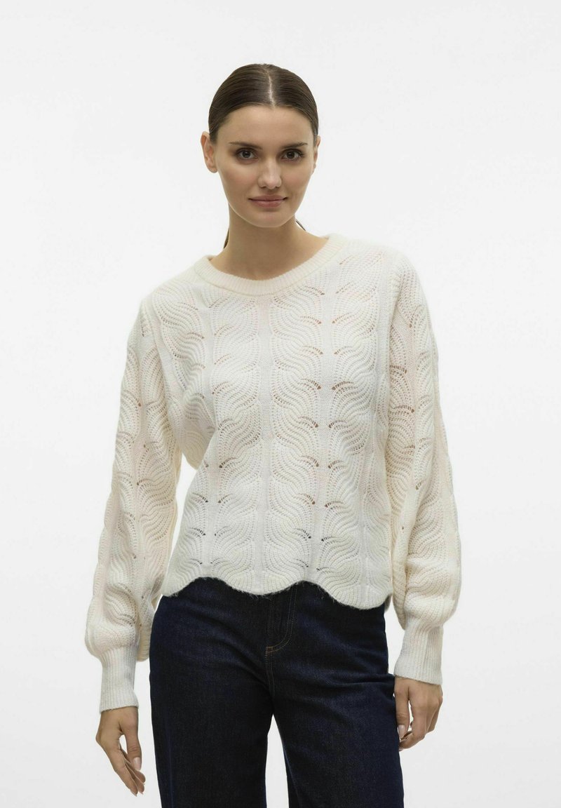 Aukštos kokybės megztinis off-white spalvos, su banguota raštų, raukiniais rankogaliais ir apvaliu hemu. Suderintas su tamsiai mėlynomis džinsomis.