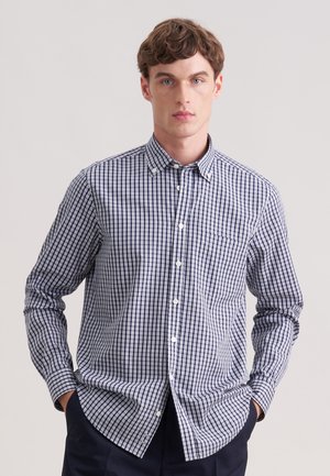 Junger Mann, der ein marineblau-weiß kariertes Hemd mit Knopfleiste und langen Ärmeln trägt, vor einem schlichten hellgrauen Hintergrund steht.