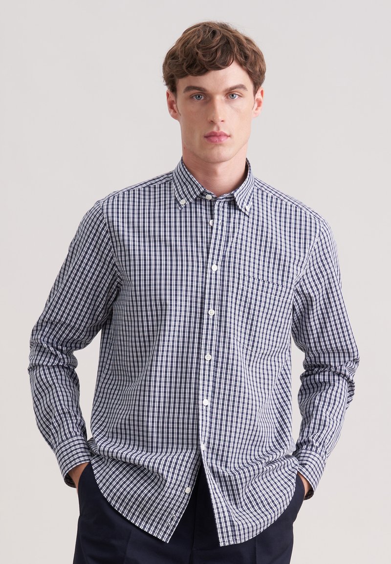 Jeune homme portant une chemise à manches longues à carreaux bleu marine et blanc, debout devant un fond uni gris clair.
