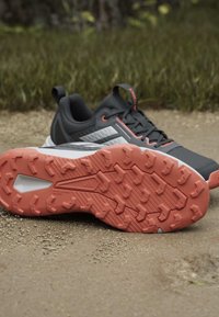 Deux chaussures de trail grises et corail avec des semelles texturées reposant sur un sentier de gravier avec de l’herbe en arrière-plan.
