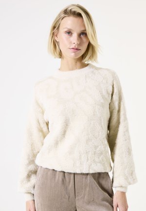 Pull en fausse fourrure de couleur crème avec des manches longues, un col rond et un motif texturé. Le tissu semble doux et moelleux. Associé à un pantalon en velours côtelé beige.