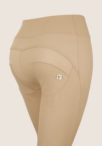 Leggings beige con una texture liscia, caratterizzati da cuciture decorative e un logo dorato nella zona della tasca posteriore.