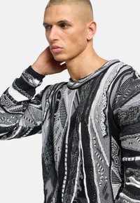 Pull en tricot à motifs noir et blanc, comprenant un mélange de textures et de designs, incluant des formes géométriques et des lignes. Encolure ronde et manches longues.
