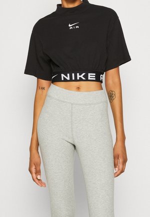 Kobieta ubrana w czarny krótki top Nike Air z logo oraz jasnoszare, wysokociśnieniowe legginsy, z widocznymi tatuażami na obu przedramionach.