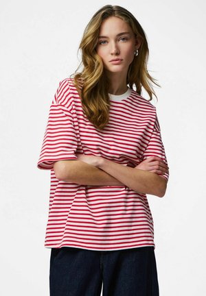 Tânără cu păr blond ondulat, purtând un tricou cu mâneci scurte roșu și alb în dungi și pantaloni închiși la culoare, cu brațele încrucișate, expresie neutră.