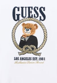 Κεντημένο έμβλημα που απεικονίζει μια αρκούδα με μαύρη και λευκή ριγωτή μπλούζα, περιβαλλόμενη από ένα χρυσό σχέδιο σχοινιού, σε λευκό φόντο με κείμενο.