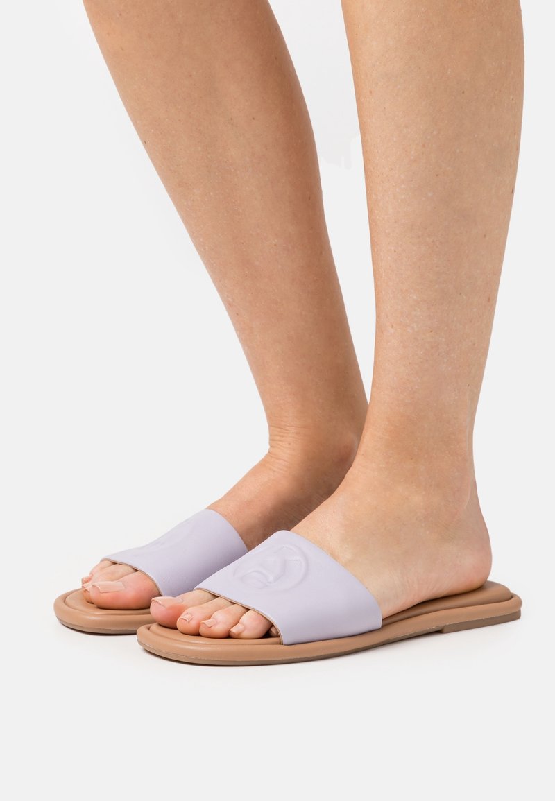 Pieds portant des sandales plates beiges à enfiler avec de larges brides violettes claires embossées d'un logo circulaire sur un fond blanc.
