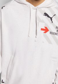 Světle šedý pulovrový svetr s tečkovanou texturou, klokaní kapsou a potisknutým logem Puma. Obsahuje výrazné grafické prvky a design šipky.